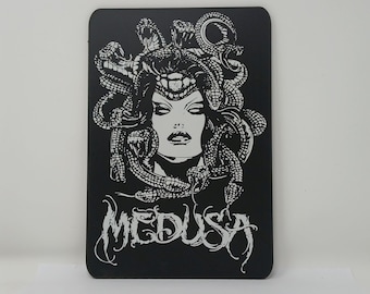 May include: Black and white illustration of Medusa, a mythical Greek figure with snakes for hair, on a black background. The text "MEDUSA" is written in a stylized font below the image.