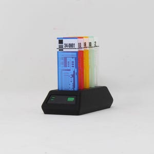 Pode incluir: Uma base preta suporta cinco objetos retangulares coloridos. Os objetos são azuis, laranja, amarelo, verde e branco. O objeto azul tem o texto "34-0901 10 19 10 2". A base tem duas luzes verdes.