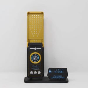 May include: A Starfleet communicator replica featuring a gold-colored flip cover with a perforated pattern, a black base with a circular display, three silver buttons, and a small black display stand labeled with the text STARFLEET COMMUNICATOR.