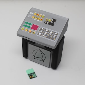May include: A gray and black console with a sloped control panel featuring yellow, red, and blue circular buttons, a numeric keypad, and text labels including NTA and STA. A black Starfleet-style delta emblem is on the front. A small green data chip lies nearby.