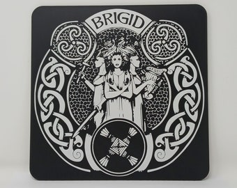 May include: Black and white coaster with a Celtic knot design and the word "BRIGID" in the center. The design features a central figure with three heads and a harp.