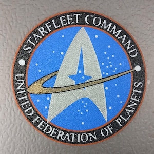 May include: Circular patch featuring the Starfleet Command emblem from Star Trek. The patch has a blue background with white stars, a silver delta, and the text "STARFLEET COMMAND UNITED FEDERATION OF PLANETS" in white lettering.