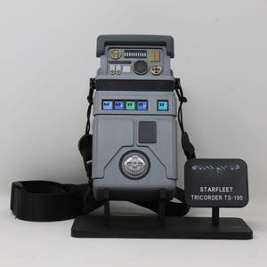 May include: A grey Starfleet Tricorder TS-190 prop replica featuring a top control panel with various buttons, a middle section with blue illuminated squares, and a circular silver dial. It includes a black shoulder strap and a display stand.