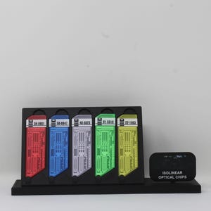 May include: A black display stand holding five rectangular isolinear optical chips in red, blue, silver, green, and yellow. Each chip features a unique serial number: 34-0901, 58-8847, 40-0023, 01-5510, and 22-1903. A small black sign reads ISOLINEAR OPTICAL CHIPS.