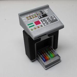 May include: A gray and black miniature control console featuring a sloped top panel with a numeric keypad, various colored indicator lights, and text labels including NTA and STA. A small drawer at the base contains several colorful rectangular inserts.