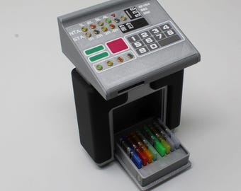 May include: A gray and black miniature control console featuring a sloped top panel with a numeric keypad, various colored indicator lights, and text labels including NTA and STA. A small drawer at the base contains several colorful rectangular inserts.