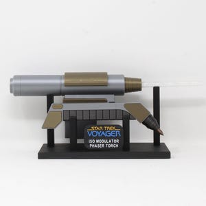Op de afbeelding: Een Star Trek Voyager ISO Modulator Phaser Torch prop replica. De prop is grijs, goud en zwart, met een heldere, gloeiende emitter. De basis heeft het Star Trek Voyager logo en tekst.