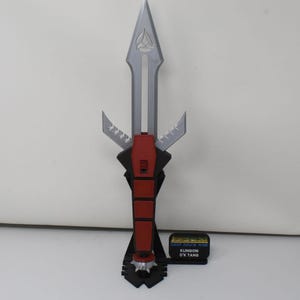 May include: A silver and red Klingon D'k tahg dagger replica mounted on a black stand. The blade features a Starfleet insignia, and a small black plaque reads DEEP SPACE NINE KLINGON D'K TAHG. The handle has a red grip and a spiked pommel.