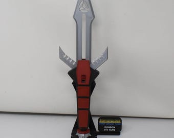 May include: A silver and red Klingon D'k tahg dagger replica mounted on a black stand. The blade features a Starfleet insignia, and a small black plaque reads DEEP SPACE NINE KLINGON D'K TAHG. The handle has a red grip and a spiked pommel.
