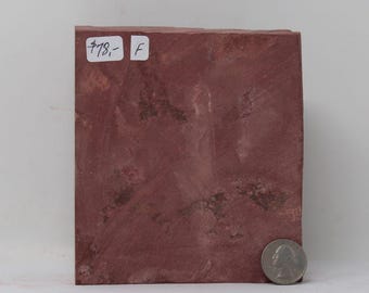 May include: A rectangular slab of reddish-brown stone with a textured, matte surface. The stone features natural dark brown inclusions and variations in color. A white sticker with the text $78,- and F is attached, and a US quarter coin is present.