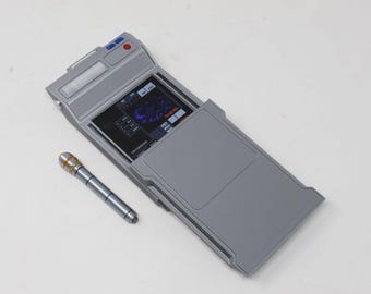 May include: A gray rectangular electronic device with a screen displaying a dark interface with blue and white text and graphics, accompanied by a small gray cylindrical stylus featuring two gold bands near the top end of the tool.