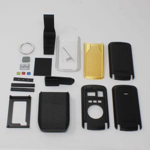 Puede incluir: Una colección de componentes electrónicos desmontados que incluye carcasas de plástico negro, un panel dorado texturizado, un marco plateado, un pequeño resorte metálico, un anillo circular y varios botones rectangulares pequeños en negro, gris, rojo, verde y azul.