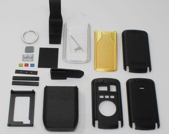 May include: A collection of disassembled electronic device components including black plastic housing shells, a textured gold panel, a silver frame, a small metal spring, a circular ring, and various small rectangular buttons in black, gray, red, green, and blue.