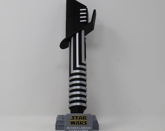 May include: A black and silver Mandalorian Darksaber replica, standing upright on a gray base. The hilt is silver with black accents, and the blade is black. The base features the Star Wars logo and the words "Mandalorian Darksaber."