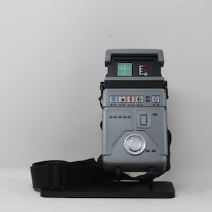 May include: A gray rectangular bag with a structured design resembling a sci-fi device. It features a top panel with a green grid display, a red light, and the letter E. The front includes blue and red square buttons, a silver circular dial, and black straps.
