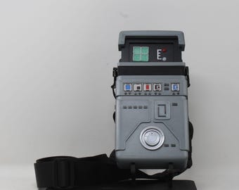 May include: A gray rectangular bag with a structured design resembling a sci-fi device. It features a top panel with a green grid display, a red light, and the letter E. The front includes blue and red square buttons, a silver circular dial, and black straps.