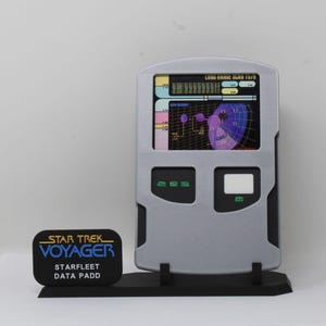 May include: A silver Star Trek Voyager data padd replica with a screen showing colourful graphics. The data padd has two rectangular buttons and a black base. A separate black plaque reads "STAR TREK VOYAGER STARFLEET DATA PADD".