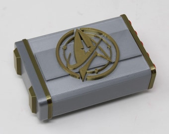 May include: A rectangular gray box with metallic gold corner accents and a central circular gold emblem featuring a stylized letter A. The box has a smooth, layered finish with four small red circular protrusions visible along one side edge.