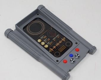 May include: A gray rectangular electronic device holder featuring a central screen display with a circular radar graphic, waveform patterns, and circular buttons. The frame includes two red buttons, one blue button, and two gray circular control pads.