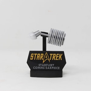 Könnte beinhalten: Eine Star Trek Starfleet Comms Earpiece Replik. Der Ohrhörer ist silber und weiß, montiert auf einem schwarzen Sockel mit dem Star Trek-Logo und Text in Gold. Das Design umfasst einen gerippten Abschnitt und einen weißen Ohrhörer.
