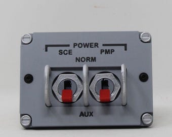 May include: A rectangular gray control panel featuring two toggle switches with red tips, protected by silver metal guards. The panel is labeled with black text reading POWER, SCE, PMP, NORM, and AUX, and is secured at the corners by four silver screws.