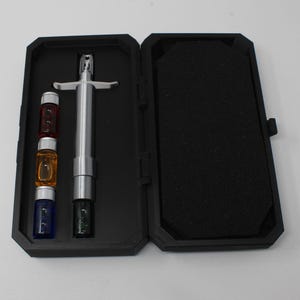 Peut inclure: Un outil en métal argenté de type seringue avec une poignée transversale, accompagné de quatre petits flacons en verre rouge, orange, bleu et noir, le tout logé dans un étui de transport rectangulaire noir doublé de mousse.