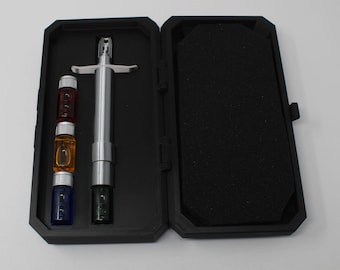 May include: A silver-colored metal syringe-style tool with a crossbar handle, accompanied by four small glass vials in red, orange, blue, and black, all housed within a black foam-lined rectangular carrying case with a hinged lid.