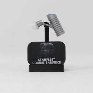 May include: A silver Starfleet comms earpiece with a white silicone tip and a ribbed cylindrical body, mounted on a black rectangular display stand featuring the white text STARFLEET COMMS EARPIECE and a small circular graphic icon.