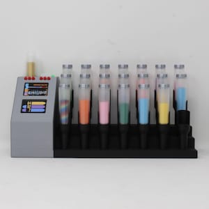 May include: A gray device with a digital display and multiple clear tubes filled with colorful sand. The tubes are arranged in a row on a black base. The sand colors include rainbow, orange, pink, green, blue, and yellow.