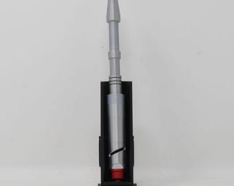 May include: A silver-colored sonic screwdriver prop with a cylindrical body, a tapered top section featuring a small red tip, and a diagonal slit detail on the lower handle, held upright in a black rectangular display stand with a flat base.