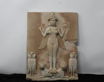 May include: A rectangular beige stone relief carving depicting a central winged figure wearing a horned headdress, standing atop two lions. The figure holds rings in raised hands, flanked by two standing owls on either side of the base.