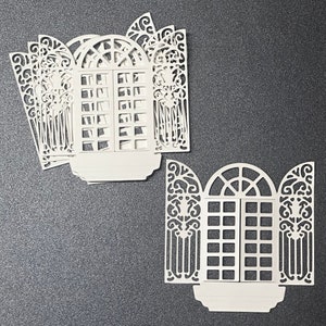 May include: Two white paper cutouts of ornate windows with arched tops and intricate designs. The windows are made of multiple layers of paper, creating a 3D effect.