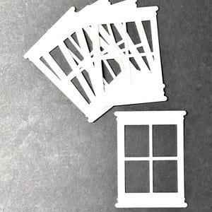 May include: White paper cutouts of miniature windows. One window is isolated, while several others are fanned out. The windows have a classic design with four panes. The background is a solid gray color.