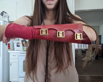 May include: A red leather arm guard with gold metal accents. The arm guard is worn on the left arm and has three buckles.