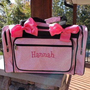 May include: Pink glitter duffel bag with black trim and two large pink bows. The bag has a zippered pocket on the front and the name "Hannah" is embroidered in white.