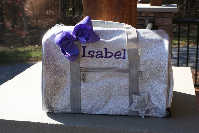 K&ouml;nnte beinhalten: Eine silberne Glitzer-Sporttasche mit einer lila Schleife und dem Namen "Isabel" bestickt auf der Vorderseite. Die Tasche hat einen Rei&szlig;verschluss und einen sternf&ouml;rmigen Aufn&auml;her.