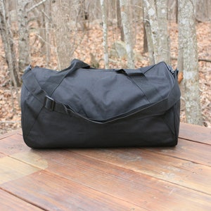 May include: A black duffel bag with a single strap and a zipper closure. The bag is sitting on a wooden surface.