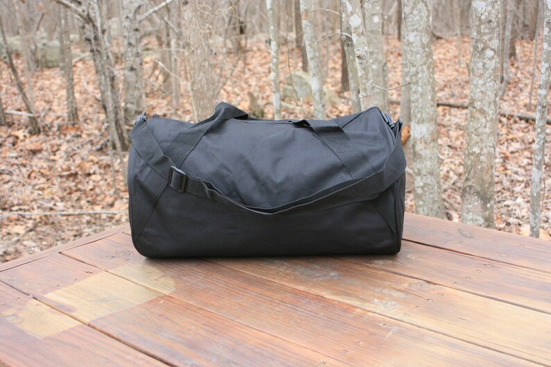 May include: A black duffel bag with a single strap and a zipper closure. The bag is sitting on a wooden surface.