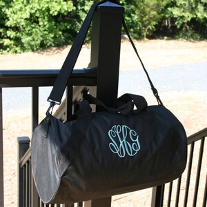 May include: A black duffel bag with a teal monogram on the front. The bag is hanging from a black strap on a black railing.