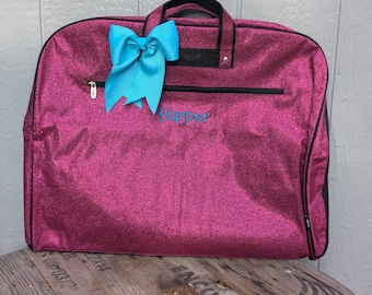 May include: A magenta garment bag with a glittery finish, a black zip, and a black handle. A bright blue bow is attached to the top. The name "Harper" is embroidered in blue on the front.