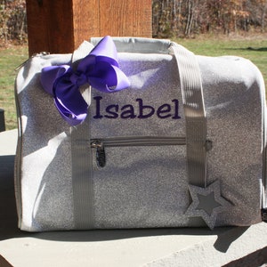 K&ouml;nnte beinhalten: Eine silberne Glitzer-Sporttasche mit einer violetten Schleife und dem Namen "Isabel" bestickt auf der Vorderseite. Ein silberner Stern ist an der Tasche befestigt.