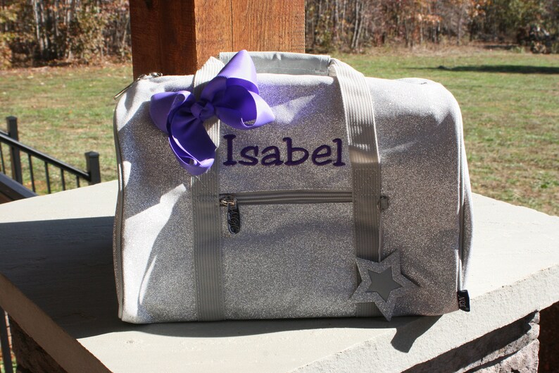K&ouml;nnte beinhalten: Eine silberne Glitzer-Sporttasche mit einer violetten Schleife und dem Namen "Isabel" bestickt auf der Vorderseite. Ein silberner Stern ist an der Tasche befestigt.
