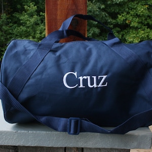 May include: Navy blue duffel bag with white embroidered text "Cruz". The bag has a shoulder strap and two handles. The bag is sitting on a stone surface.