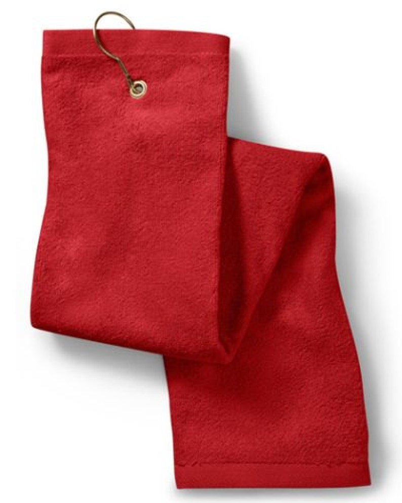 Puede incluir: Una toalla de golf rectangular de color rojo s&oacute;lido con una superficie de tela texturizada. La toalla incluye un ojal de metal color lat&oacute;n en la parte superior central, equipado con un gancho de metal a juego para colgarla.