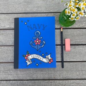 May include: A blue journal with a navy anchor and the word "NAVY" in white lettering. The anchor has a red flower and a white ribbon with the word "JOURNAL" on it. The background is blue with white stars.