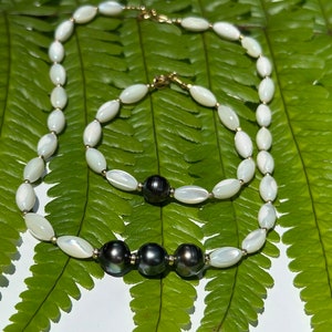 May include: Two necklaces with white oval beads and black pearls. The necklaces are on a green fern leaf background.