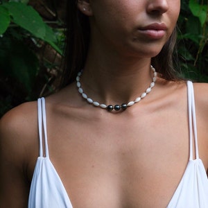 Op de afbeelding: Een vrouw draagt een witte bikini top met een witte en zwarte parelketting. De ketting heeft drie zwarte parels in het midden.