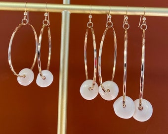 May include: A display of gold-colored hoop earrings with white circular pendants. The earrings vary in size and shape, some with a hammered texture. The earrings are hanging on a gold-colored display stand against a brown background.