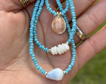 May include: A turquoise beaded necklace with two shell pendants. The necklace is layered and has a delicate gold chain.