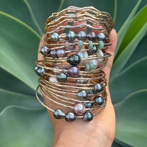 May include: A stack of gold-toned bangles, some with a hammered texture, adorned with dark gray, lavender, and white pearls, and small green beads. The bracelets are stacked on top of each other, creating a layered effect.